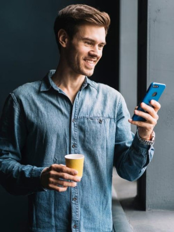 Smiling young man holding takeaway coffee cup looking cellphone 1